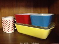 View of white ceramic mug with red heart pattern next to Pyrex Fridgie containers in red, blue, and yellow with glass lid on shelf.