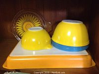 Photo of three Pyrex bowls including a sunflower-patterned clear glass bowl, two yellow mixing bowls (one smaller with spout, one larger with blue base), and an orange and white rectangular lasagna/baking pan with clear lid.