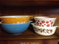 Four vintage glass mixing bowls stacked on shelf: two large Pyrex (blue and orange with floral), one medium Pyrex white with red polka dots, one smaller Federal bowl with brown floral design
