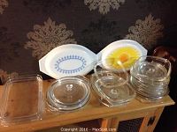 Photo showing multiple glass Pyrex lids stacked and ceramic Corningware lids with flower and blue patterns displayed on top of a wooden surface.