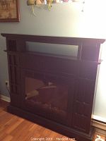 Dark wood electric fireplace with large glass front and top display shelf, shown plugged in against a wall with hardwood floor.