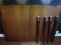 Table top resting upright showing its solid wood finish and rounded corners along with four detached wooden legs placed upright against it.