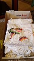 Box of crocheted table cloths and pillow covers including a pillow cover with a colorful dragon embroidery partially covering white crocheted pieces