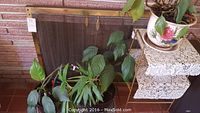 Two metal fireplace frames with mesh screens, one metal table with a white lace cover, and two potted leafy plants in decorative ceramic pots