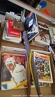 Photo showing several stacked sports magazines including Breakaway and CFL magazines, a closed umbrella lying on top of the magazines, lamp base visible with power cord, and other sports books.