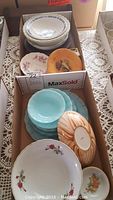 Box with stacked white plates with blue detailing, orange marbled tray, turquoise bowls and plates, and white plates with floral prints.