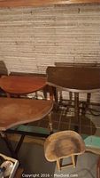 Four wooden tables arranged along a wall with textured wallpaper and tile floor. Different shapes and sizes visible, including a stool in front.