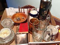 Box containing assorted vintage barware items including mugs, ashtrays, and metal framed lamp