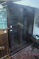 Full view of the antique wooden linen press cabinet showing the dark wood, glass panel double doors with interior shelves, and ornate details.