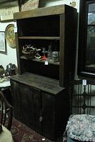 Photo of early Long Island wooden hutch showing full front with three tiers and single door lower cabinet. Hutch is dark wood with open shelves on top and closed cabinet door below. Surrounded by other furniture and items with visible price tag.