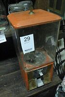 Full view of the vintage gumball/candy dispenser showing upright rectangular shape with orange metal base and clear plastic container, top lid with wear.