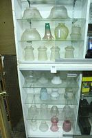 Full display cabinet showing multiple shelves with various glass lamp shades of different sizes, colors and designs including frosted and colored glass shades.