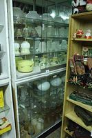 Wide view of glass cabinet shelves holding multiple different lampshades including clear, white, ceramic, and brass.