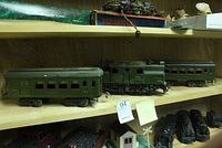 Three vintage green metal train cars on a wooden shelf, showing engine, passenger car, and caboose.