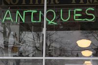 Green neon tube spelling 'ANTIQU' and white neon tube spelling 'ES' forming 'ANTIQUES' in a window display.