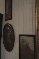 Photo showing two framed wall hangings on a white wooden wall; one rectangular landscape painting in a black frame and an oval framed photograph with domed glass.