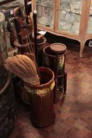 Photo of two pottery umbrella stands, one brown ribbed with beige top, another green floral relief, and carved wooden umbrella stand with umbrellas and broom inside.