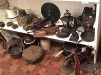 Shows the entire collection arranged on white shelves and floor, featuring jugs, pots, pans, metal tools, and lanterns.