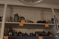 Shelves showing a collection of various pewter coffee and tea pots, creamers, sugar bowls, and other containers. Items are displayed in two rows on wooden shelving.