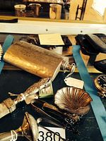 Photo shows a display of vintage silver items including a hammered silver-plated flask, silver mesh purse, several baby rattles, silver tongs, a small silver dish, and a pocket watch on a blue cloth.