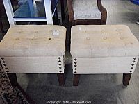 Two beige upholstered tufted ottomans shown side-by-side, square shape, wooden tapered legs, bronze-tone nail head trim visible on corners.