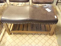 Front view of wooden bench showing dark brown leather cushioned top with nail head trim and paneled bottom shelf made of wooden slats.