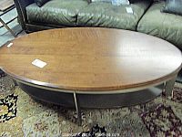 Oval wooden coffee table with metal frame and wooden shelf underneath, sitting on a patterned rug with some wear and light scratches visible on the top.