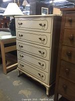 Front and side view of the vintage white dresser showing all five drawers with gold-toned handles and accents, and curved leg base on caster wheels.