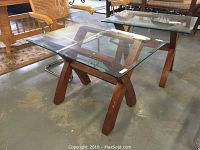 Side view of square beveled glass side table with crossed wooden legs on a concrete floor showing overall condition and design