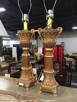 Pair of matching lamps with gold accents and distressed tan body standing on floor in warehouse setting.