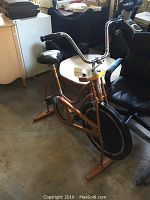 Side angle view of the vintage metallic orange Schwinn stationary exercise bike showing seat, frame, and front black wheel cover.