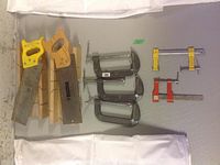 Photo showing two hand miter saws with yellow and wooden handles, two wooden miter guides, and multiple C-clamps in various sizes and types on a grey surface.