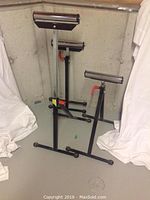 Three black metal roller stands with red adjustment knobs and horizontal metal rollers, set on basement floor.