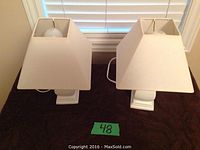 Front view of two bedside lamps with white ceramic bases and matching white square shades placed on a brown surface in front of a window with blinds.