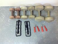 Photo showing all items in the lot arranged on a grey floor: two pairs of beige dumbbells (10 lb and 5 lb), one pair of brown 2.5 lb dumbbells, two black wrist weights, and two orange spring hand/forearm builders.