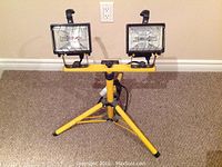 Front view of dual quartz halogen work lights on yellow tripod stand, showing both lamps and tripod base on carpeted floor against a beige wall.