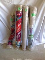 Four rolls of fabric standing upright against a white background; two wrapped burlap jute rolls with red and blue packaging and two beige garden cloth rolls.
