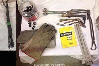 Welding gear including torches, gloves, welding rods, torch tips, jar of autobody clips, and welding manual laid out on white cloth.