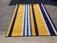 Full view of the rectangular area rug laid flat on pavement showing alternating stripes in mustard yellow, navy blue, light blue, and white.