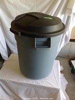 Blue Rubbermaid Roughneck garbage can with black lid shown on white table in storage area.