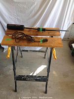 Folding work table with wooden plank top, black metal legs, and yellow clamp handles, showing wear on wood components.