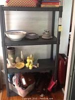 4-tier black plastic shelving unit with various small household items stored on shelves, including plates, bowls, decorative items, and a suitcase beside it.