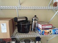 Shelf with small Crock Pot, French press-style hot chocolate maker, box with cookie press, multicolored mugs, and cardboard box.