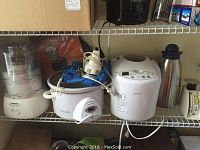 Photo showing kitchen appliances including Cuisinart, Crock-Pot, Sunbeam appliances and a metal vacuum flask on a shelf.