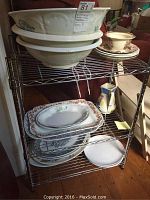 Metal shelving rack holding the dishes: three large white basins with floral transferware patterns stacked on the top shelf; below are several rectangular and oval platters and dinner plates with floral and scenic designs, and a ceramic pitcher with blue and brown geometric painting on the side.
