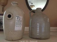 Two stoneware jugs are displayed side by side. One jug is round with a hole in the middle and stamped maker's mark. The other jug is plain with handle and spout. Both are light beige to gray in color with glazed finish.