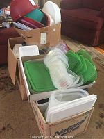 Boxes filled with over 20 plastic food storage containers with mostly green lids and some white lids. Containers are clear plastic and stackable.