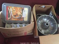 Two cardboard boxes containing various metal bakeware including bundt pans, muffin tins, and a vintage pie tin with a Table Talk label visible on the lid.