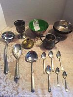 Photo showing all items: two silver bowls (one green enameled), brass candlestick, goblet, six souvenir spoons, and three large serving spoons on a beige lace tablecloth.