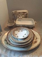 Photo showing stacked plates, lidded casserole dish, cup and saucer, and serving platter propped behind, all in brown and white transferware style.